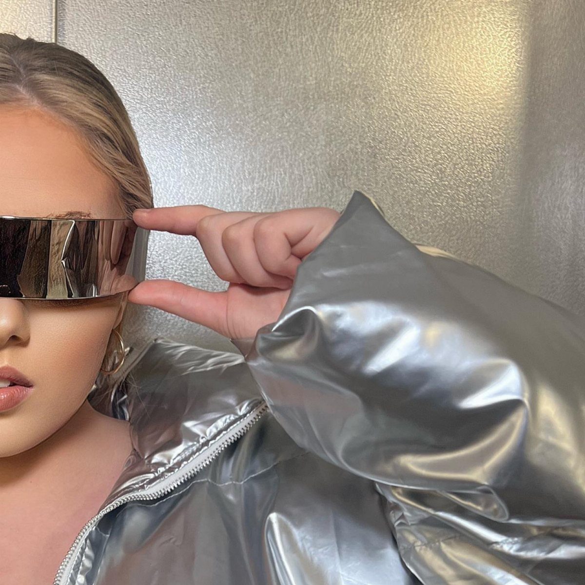 A person wearing silver shield glasses and a metallic shiny silver jacket against a silver wall.