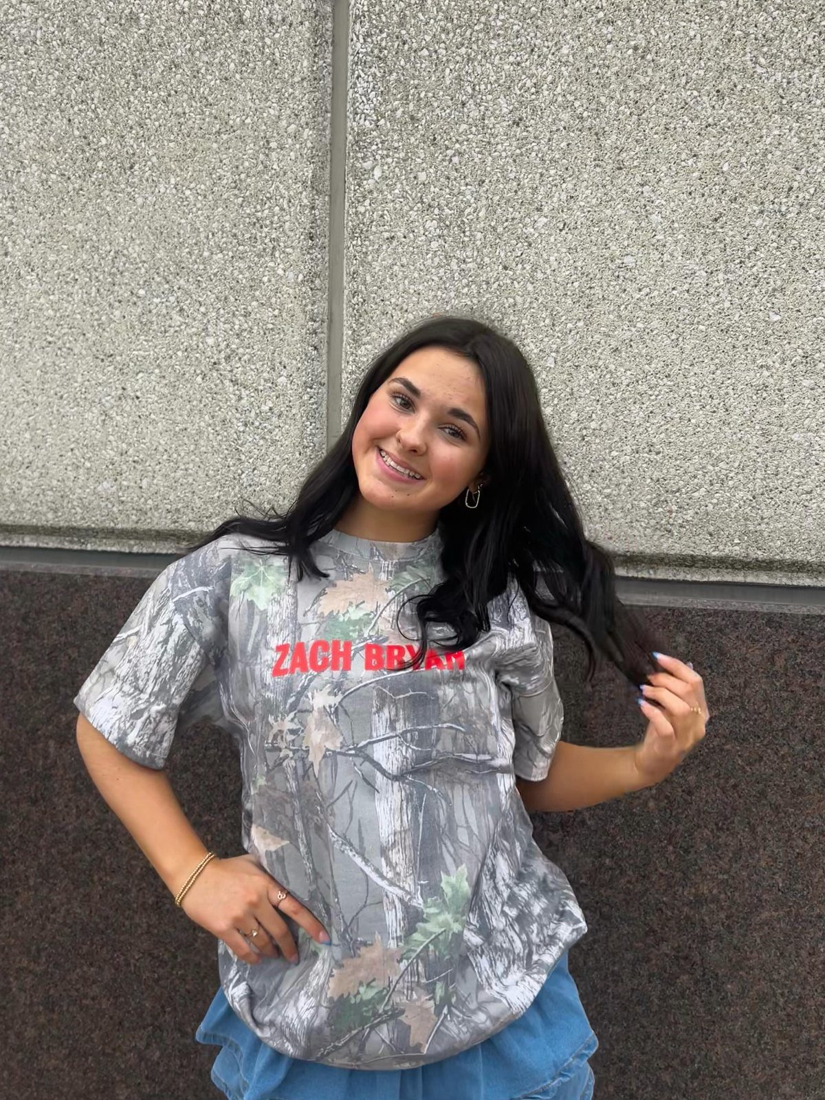 A smiling woman wearing a Zach Bryan camouflage t-shirt and blue denim shorts against a grey wall.