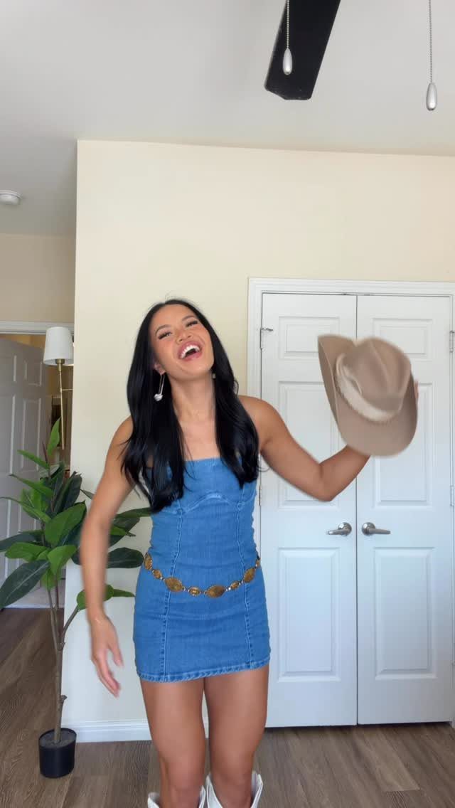 A smiling woman in a blue denim mini dress holding a tan cowboy hat indoors.