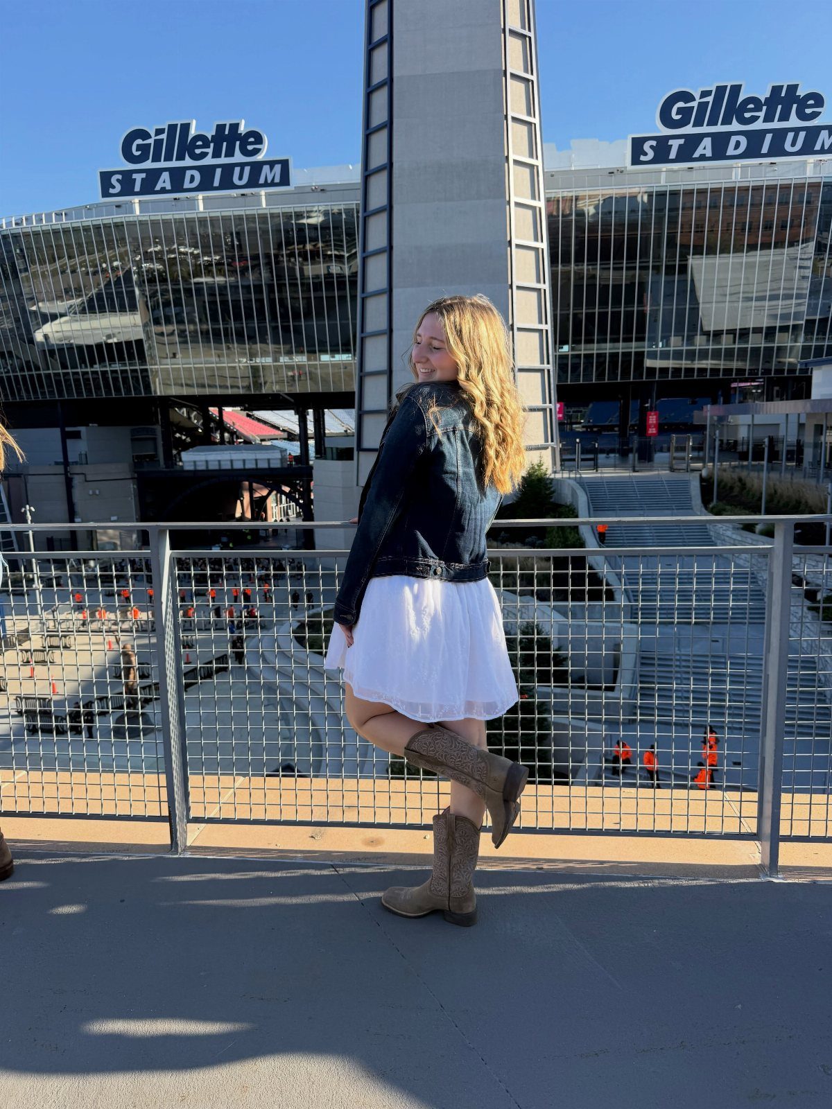 A woman wearing a black denim jacket, white skirt, and cowboy boots posing near a stadium.