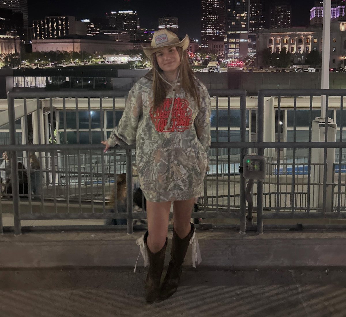 A young woman wearing a cowboy hat, patterned hoodie, and fringed cowboy boots at night.