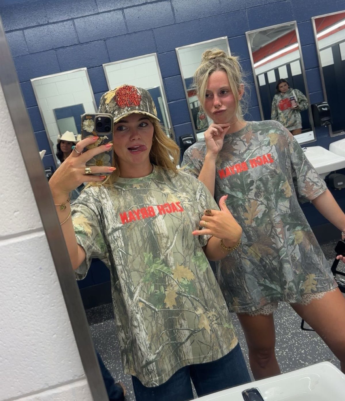 Two women posing in a mirror wearing matching camouflage graphic t-shirts and a camouflage hat.
